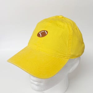 Op de afbeelding: Een felgele baseballcap met een gebogen klep. De cap heeft een geborduurd bruin American football-ontwerp op de voorkant. De cap is gemaakt van een zachte stof en heeft een knoop aan de bovenkant.