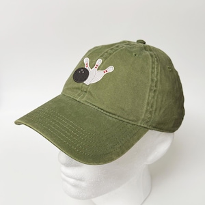 May include: An olive green baseball cap featuring an embroidered graphic on the front panel. The design depicts a black bowling ball with three white finger holes positioned next to three white bowling pins with red stripes near the top.