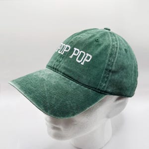 May include: A green baseball cap with the embroidered text "POP POP" in white. The cap has a curved brim and a distressed, vintage look. The hat is displayed on a white mannequin head.