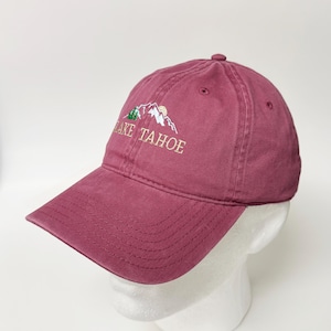Puede incluir: Gorra de béisbol granate con el texto bordado "LAKE TAHOE" en hilo dorado. El diseño incluye una cordillera y árboles. La gorra tiene una visera curva.