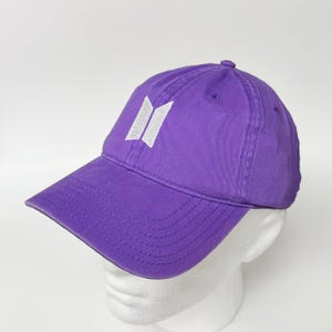 Peut inclure: Casquette de baseball violette avec une visière incurvée. Le chapeau présente un logo blanc brodé sur le devant. La casquette est en tissu doux et a un design classique à six panneaux.