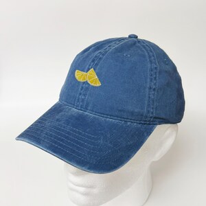 Pode incluir: Boné de beisebol azul denim com aba curva. O boné apresenta um design bordado de duas fatias de limão amarelas na frente. O chapéu é exibido em uma cabeça de manequim branca.