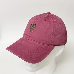Könnte beinhalten: Eine weinrote Baseballkappe mit gebogenem Schirm. Die Kappe hat ein kleines, gesticktes grünes Baumdesign auf der Vorderseite. Die Kappe besteht aus einem weichen, strukturierten Stoff und hat ein klassisches Sechs-Panel-Design.