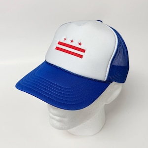 Może przedstawiać: Niebiesko-biała czapka typu trucker z emblematem flagi Waszyngtonu, D.C. Czapka ma biały panel przedni z czerwonym emblematem, niebieski daszek i niebieski tył z siatki. Czapka jest prezentowana na białej głowie manekina.