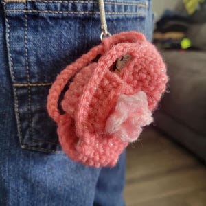 May include: A pink crocheted purse with a flower detail hanging from a pair of blue jeans.