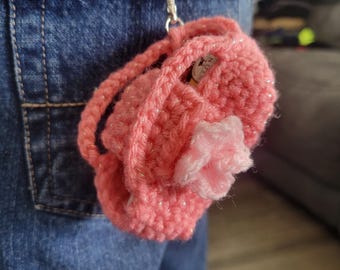 May include: A pink crocheted purse with a flower detail hanging from a pair of blue jeans.