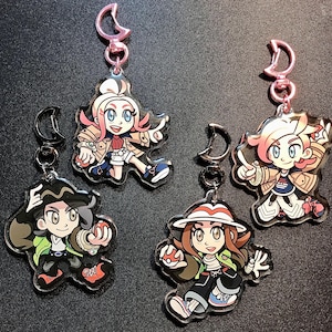 May include: Four character keyrings with pink and silver clasps. Each keyring features a different cartoon character with distinct outfits and hairstyles. The characters are in dynamic poses, holding objects.