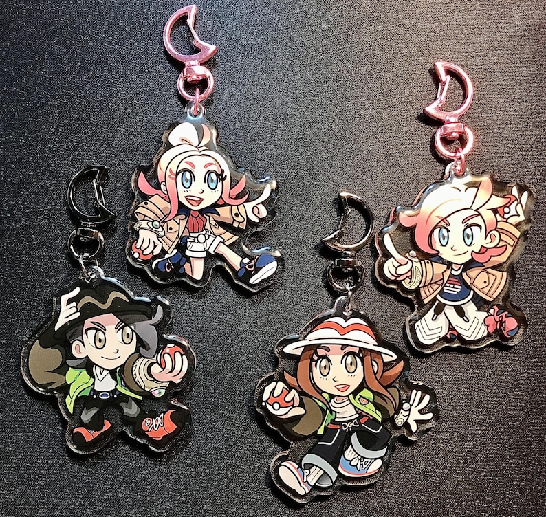 May include: Four character keyrings with pink and silver clasps. Each keyring features a different cartoon character with distinct outfits and hairstyles. The characters are in dynamic poses, holding objects.