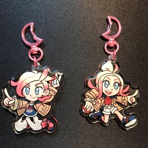 May include: Two anime-style keyrings featuring characters with pink hair, blue eyes, and tan jackets. Each keyring has a pink clasp. One character is pointing, and the other is holding a small ball.
