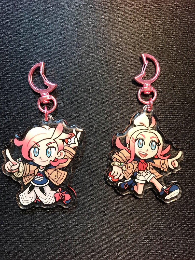 May include: Two anime-style keyrings featuring characters with pink hair, blue eyes, and tan jackets. Each keyring has a pink clasp. One character is pointing, and the other is holding a small ball.