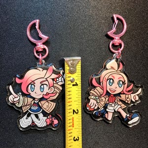 May include: Two cartoon character keyrings with pink hair and pink key rings. The keyrings are approximately 7.6 cm tall. The characters are wearing jackets, shorts, and trainers. One character is pointing, and the other is in a running pose.