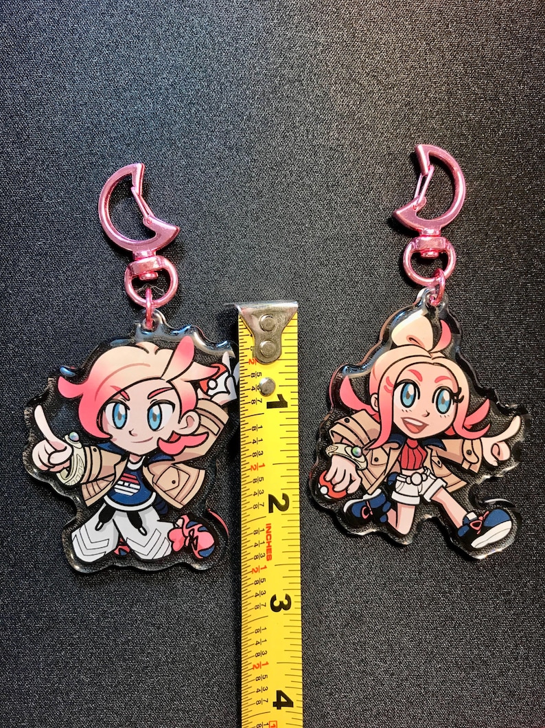 May include: Two cartoon character keyrings with pink hair and pink key rings. The keyrings are approximately 7.6 cm tall. The characters are wearing jackets, shorts, and trainers. One character is pointing, and the other is in a running pose.