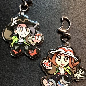 May include: Two acrylic keychains with cartoon character designs. One shows a character with black hair, a green jacket, and red shoes. The other depicts a character with brown hair, a white hat, and a red and white striped brim.