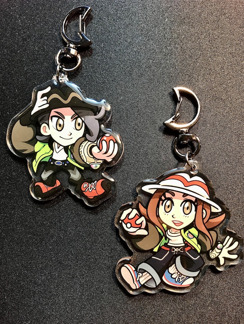 May include: Two acrylic keychains with cartoon character designs. One shows a character with black hair, a green jacket, and red shoes. The other depicts a character with brown hair, a white hat, and a red and white striped brim.