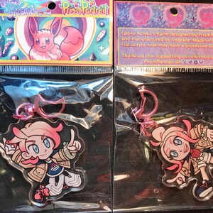 May include: Two clear acrylic charms featuring anime-style characters with pink hair and blue eyes, packaged in clear plastic. The charms have pink metal clasps. The packaging includes text that reads "Prism Portrait".