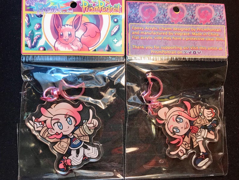 May include: Two clear acrylic charms featuring anime-style characters with pink hair and blue eyes, packaged in clear plastic. The charms have pink metal clasps. The packaging includes text that reads "Prism Portrait".