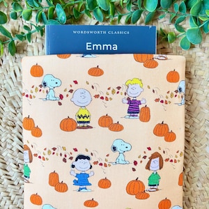 May include: A peach-coloured fabric book sleeve featuring a repeating pattern of Peanuts characters including Snoopy, Charlie Brown, and Lucy surrounded by orange pumpkins and autumn leaves, with a blue Wordsworth Classics Emma book inserted inside.
