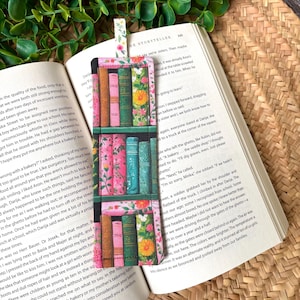 May include: A rectangular fabric bookmark featuring a printed pattern of bookshelves filled with colourful books and floral accents. The bookmark has a small fabric tab at the top with a white background and a delicate pink and green floral print.