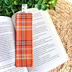 Peut inclure: Un marque-page rectangulaire en tissu avec un motif à carreaux orange, marron et blanc. Une petite languette blanche dépasse du haut, ornée d'un imprimé répétitif de petites citrouilles de différentes couleurs, dont vert, orange et marron.