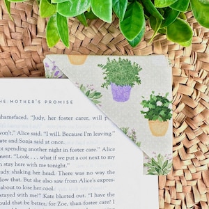 May include: A fabric corner bookmark with a botanical design of potted plants in purple, yellow, and green on a light beige backdrop. The bookmark is on a book corner with text visible. The background includes woven basket material and green leaves.