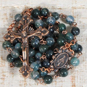 May include: A rosary with dark green and blue beads. The rosary features a copper-colored crucifix and a small oval pendant. The beads are strung together with copper-colored chain links. The rosary is a religious item.