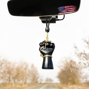 May include: A black fist car charm hanging from a rearview mirror. The charm is detailed with gold-colored wire and a gold-colored ring. The rearview mirror reflects a faded American flag.