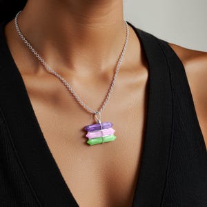 May include: A silver chain necklace with a pendant featuring three stacked crystal-shaped stones in purple, pink, and green. The crystals are wrapped with silver wire. The necklace is displayed on a person wearing a black top.