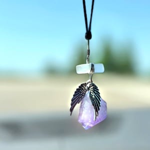 May include: A silver pendant with a purple amethyst crystal and angel wings hangs from a black cord. The pendant is a unique and stylish accessory.