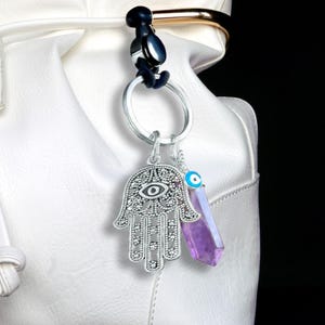 May include: A silver keychain with a Hamsa hand charm, an evil eye charm, and a purple crystal pendant. The keychain is attached to a white purse with a black background.