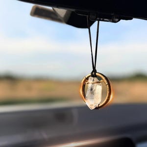 May include: A clear quartz crystal is suspended from a black string and encased in a gold wire circle. The crystal is hanging from the rearview mirror of a car.
