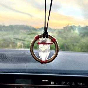 May include: A clear quartz crystal point is suspended in a silver ring with red wire wrapped around it. The crystal is hanging from a black string from the rearview mirror of a car. The car's dashboard is visible in the background with the text "Preconditioning/Ventilation Start now" displayed on the screen.