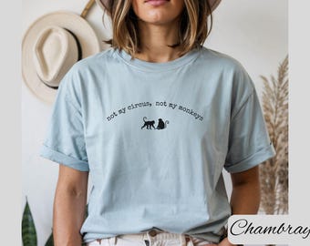 May include: Light blue t-shirt with the text "not my circus, not my monkeys" in a curved font. Two black monkey silhouettes are centered below the text. The shirt is a solid color and has rolled-up sleeves.