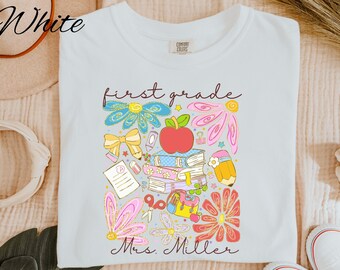 May include: White t-shirt with a colorful graphic design featuring the words "first grade" and "Mrs. Miller". The design includes an apple, books, flowers, a pencil, and other school-related elements. The shirt is a Comfort Colors brand.