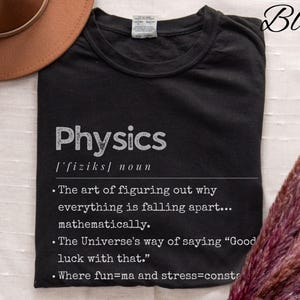Könnte beinhalten: Schwarzes T-Shirt mit dem Wort "Physics" in Weiß, zusammen mit einer Definition und humorvollen Aussagen über Physik. Ein brauner Hut und eine dekorative Pflanze befinden sich im Hintergrund.