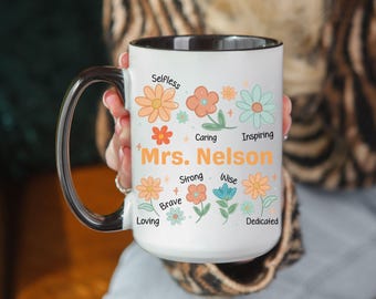 Op de afbeelding: Witte keramische mok met een zwart handvat en rand, versierd met kleurrijke bloemenillustraties en de naam "Mrs. Nelson" in oranje. De mok is versierd met woorden als "Selfless", "Caring" en "Inspiring".