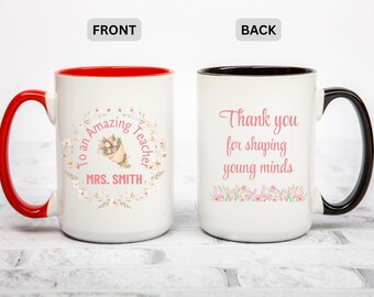 Op de afbeelding: Witte keramische mok met een rode handgreep en binnenkant. De voorkant heeft de tekst "To an Amazing Teacher" en "Mrs. Smith" met een bloemenontwerp. De achterkant zegt "Thank you for shaping young minds."