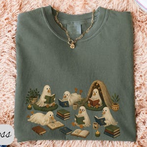 May include: A moss green crewneck sweatshirt featuring a graphic print of several white ghost-like figures reading books. The ghosts are depicted in various poses, some sitting on cushions, others under a small blanket fort, surrounded by stacks of books.