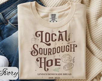 May include: Ivory-colored t-shirt with the words "Local Sourdough Hoe" in brown script, along with illustrations of bread and a jar of honey. The text "Linda's Homemade Bread EST. 2023" is also printed on the shirt.