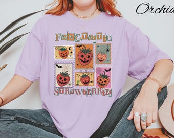 Pode incluir: Uma camiseta lilás clara com um design de Halloween. A camisa apresenta seis gráficos em estilo selo postal de morangos com rostos de jack-o'-lantern, morcegos e teias de aranha. O texto "FANTASTIC STRAWBERRIES" é impresso.