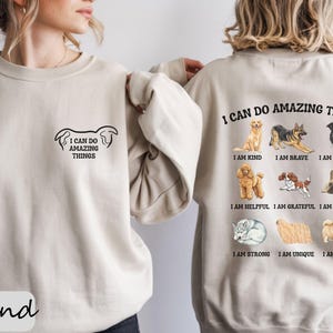 May include: A sand-coloured sweatshirt featuring the phrase "I CAN DO AMAZING THINGS" and dog illustrations. The front has a small design, and the back displays various dog breeds with affirmations such as "I am kind" and "I am brave."