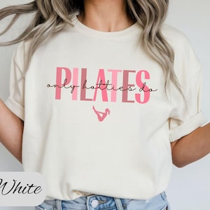 May include: Cream-colored t-shirt with the text "PILATES only hotties do" in pink. The shirt features a small pink graphic of a person doing Pilates. The word "White" is written in cursive at the bottom.