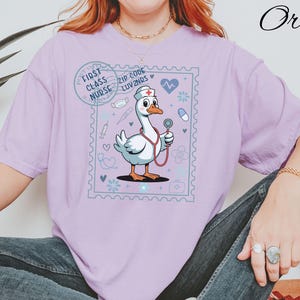 May include: A light purple t-shirt featuring a cartoon goose dressed as a nurse, holding a stethoscope. The design is framed within a postage stamp border and includes the text "First Class Nurse" and "Zip Code Luv2Nrs".