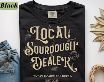 May include: A black t-shirt featuring the cream-colored text "LOCAL SOURDOUGH DEALER" in a decorative font, accompanied by illustrations of a jar, a spoon, wheat stalks, and a loaf of bread. Below, the text reads "LINDA'S HOMEMADE BREAD EST. 2023".