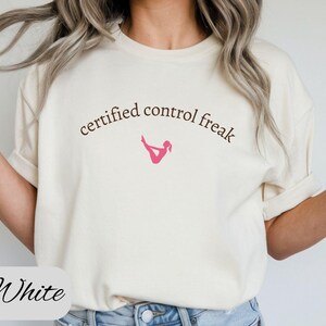 May include: Cream-colored t-shirt with the text "certified control freak" in a curved, dark brown font. A pink graphic of a person in a yoga pose is below the text. The word "White" is written in cursive at the bottom.