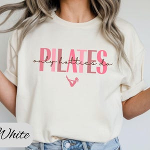 May include: Cream-colored t-shirt with the text "PILATES only hotties do" in pink. The shirt features a small pink graphic of a person in a pilates pose. The word "White" is written in cursive at the bottom.