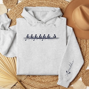 Puede incluir: Sudadera con capucha gris claro con un diseño de equipo de remo azul marino en la parte delantera. La manga tiene el nombre "Maddie" en cursiva con remos cruzados. La sudadera tiene una capucha con cordón y un bolsillo de canguro.