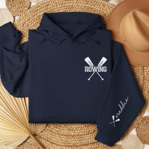 May include: Navy blue hoodie with a white "ROWING" graphic and crossed oars on the chest. The sleeve features the name "maddie" in white script. The hoodie has a hood and long sleeves.