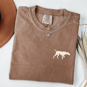 May include: A brown Comfort Colors t-shirt with a small cream-colored dog graphic. The shirt has a crew neck and short sleeves. A silver necklace with a pendant is draped across the shirt.