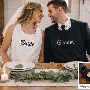 May include: A couple at a wedding reception wearing bibs. The bride's bib is white with "Bride" embroidered in black. The groom's bib is black with "Groom" in white. The table is set with candles, plates, and greenery.