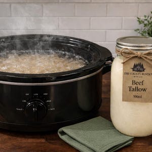 Op de afbeelding: Een zwarte slowcooker met stomende inhoud staat naast een pot rundertalg. De pot heeft een label met de tekst "Beef Tallow 500ml". Een groene doek staat op de voorgrond.